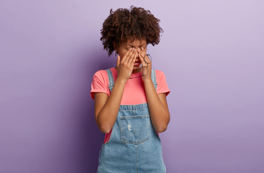Young woman standing against a purple background as she rubs her eyes.