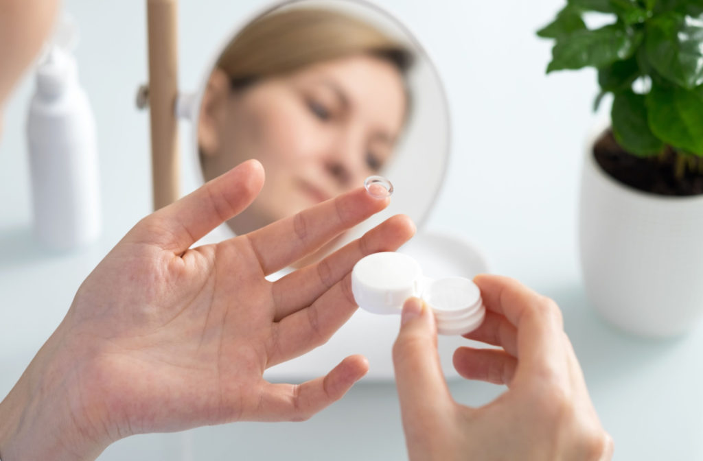 A woman holding a contact lens and case with her reflection showing in the mirror.