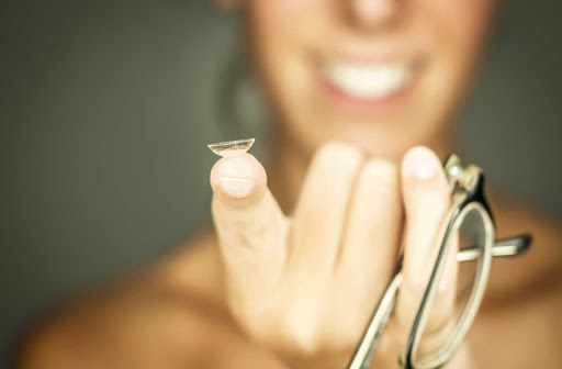 A close up of a contact lens sitting on a woman's finger while she holds a pair of glasses.