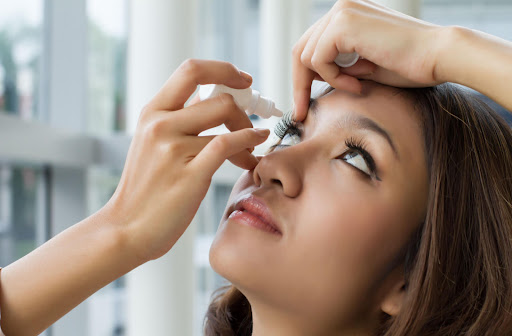 A lady is holding a small white bottle near to right eye and pouring drops of atropine fro myopia control.