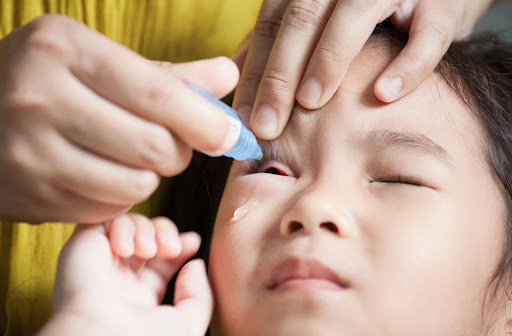 A parent lays her child down and puts eye drops on the lower eyelids of the child.