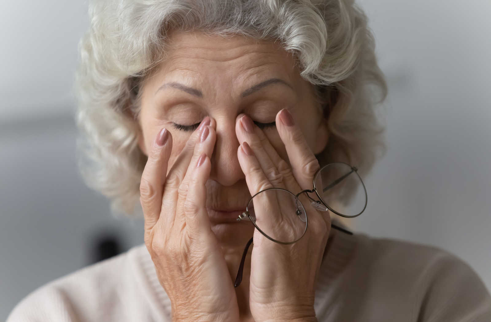 An elderly woman removes her eyeglasses and rubs her nose bridge. dry eye disease
