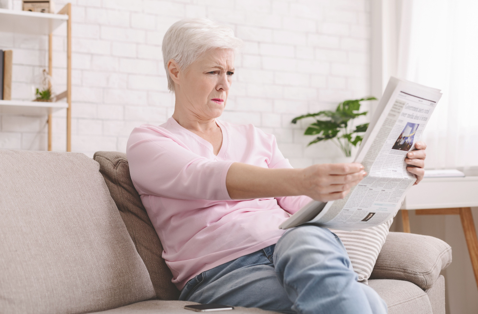 Mature woman holding a newspaper further away from herself to be able to read it.
