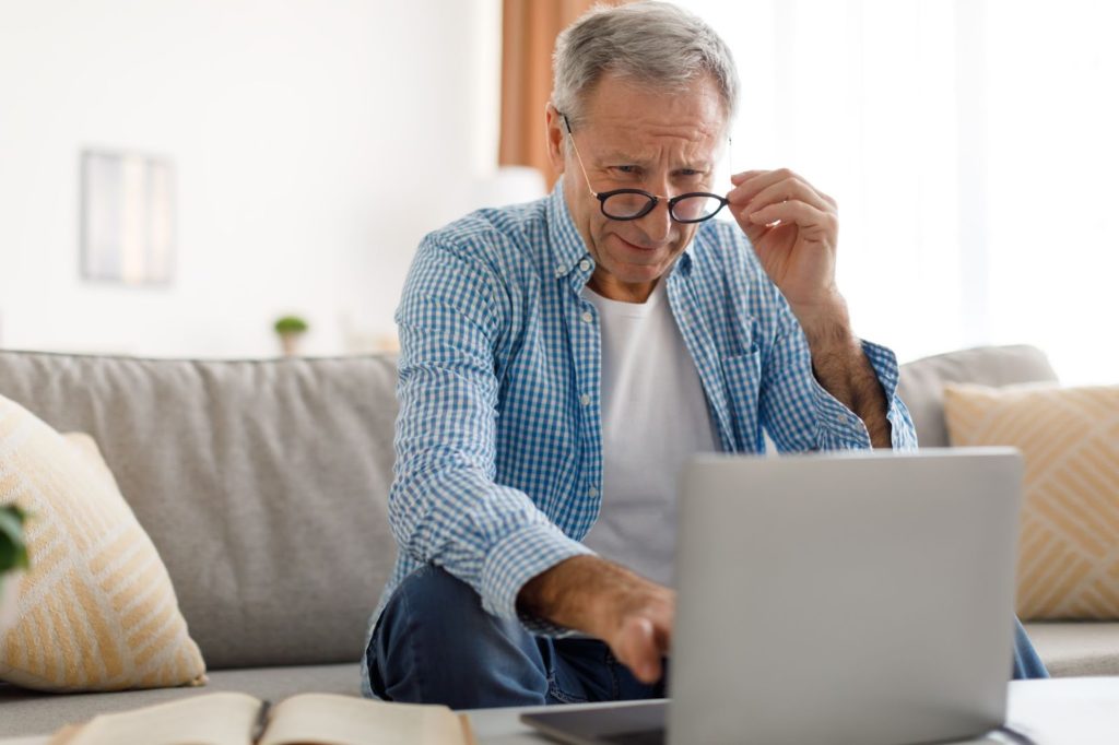 An older adult sitting on a couch and adjusting his reading glasses as he squints to read something on his laptop screen.