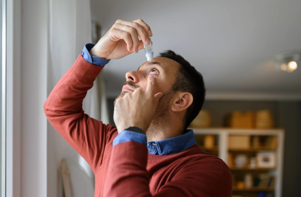 A person standing in front of a window at home and looking up as they apply eye drops to their left eye.