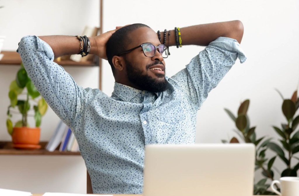 A person sitting in front of a laptop with their arms raised behind their head, taking a break to look away from their laptop and rest their eyes.