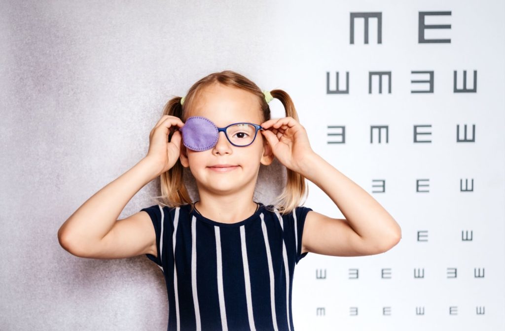 Young girl with glasses and a purple eye patch reaching up to adjust her glasses as she stands in front of a grey tumbling E eye chart.