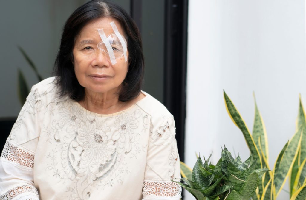 A patient relaxes in a brightly lit room, seated beside green plants, with a gentle smile on their face. A translucent eye patch covers their left eye as they recover from retinal detachment surgery.