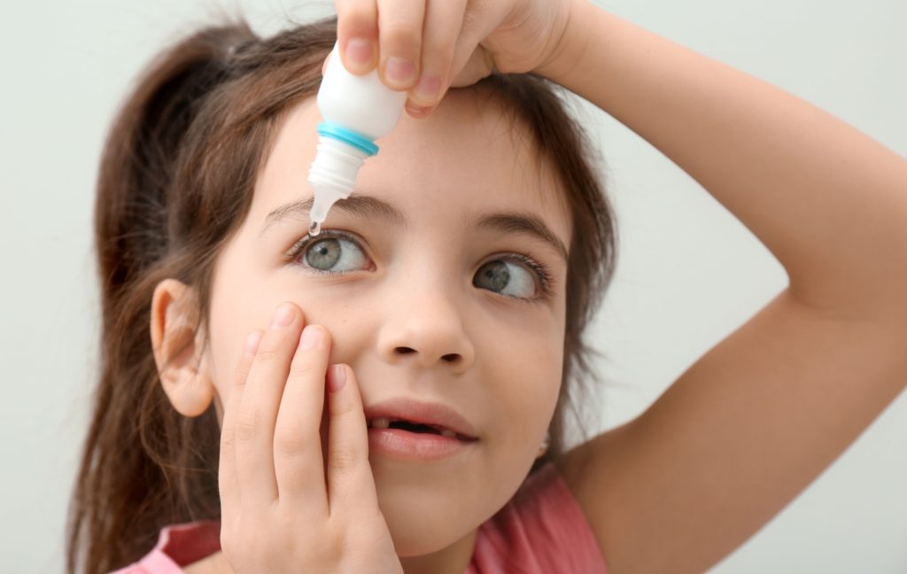 A young child gently squeezes some low-dose atropine eye drops into their left eye to help prevent myopia progression.