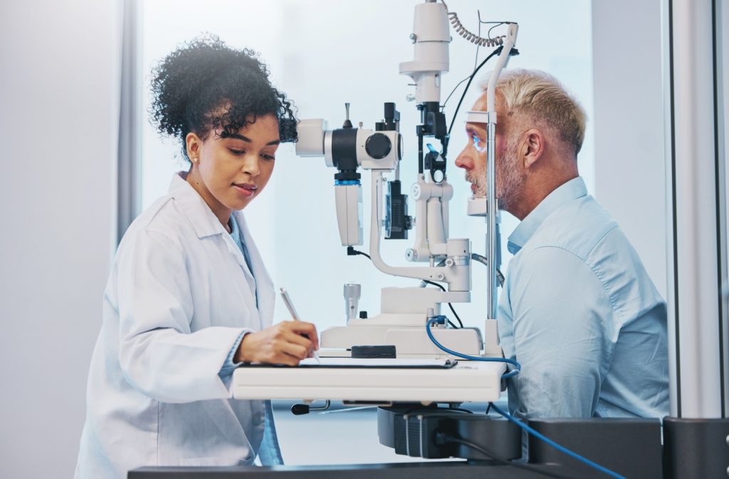 An optometrist in a white coat takes notes while examining a patient using a slit lamp in an eye clinic.