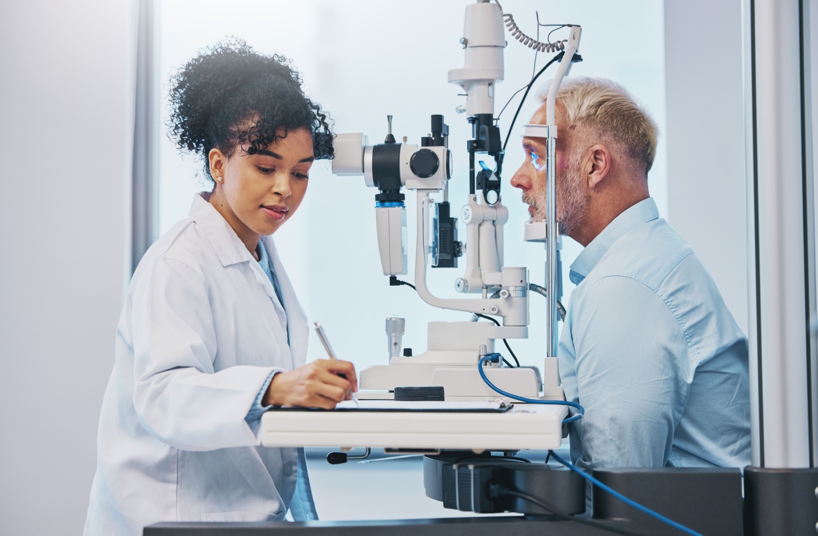 An optometrist in a white coat takes notes while examining a patient using a slit lamp in an eye clinic.