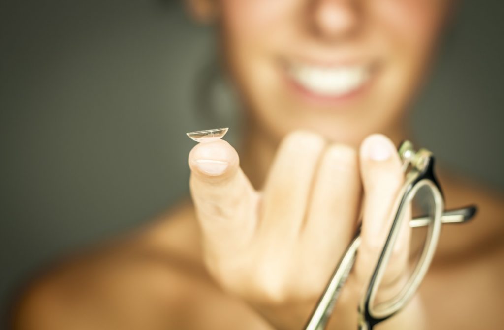 A close-up image of a hand with a contact lens on the index finger tip and eyeglasses hanging from the opposite pinky finger.