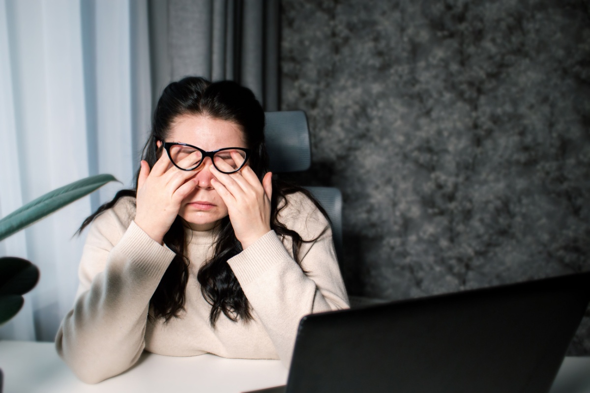 Woman wearing glasses rubbing her eyes while sitting at a desk in front of a laptop indoors.