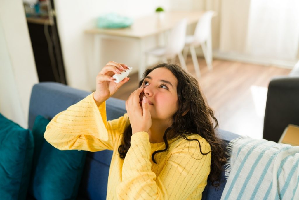 Young woman sitting on a couch applying eye drops to her eye in a bright living room.