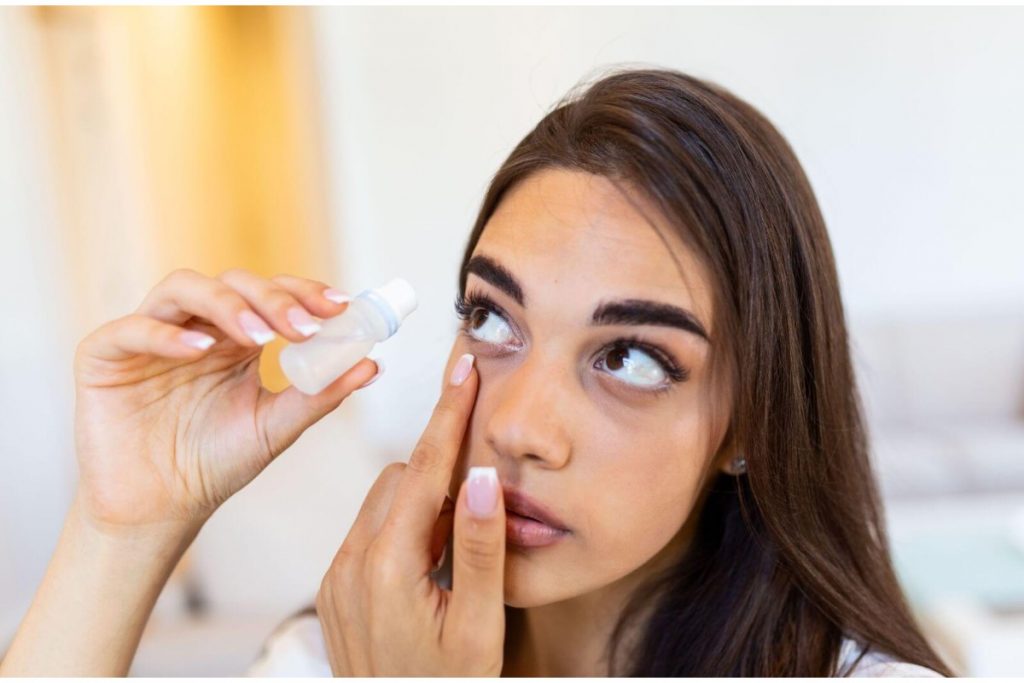A close-up of a woman with long brown hair holding down her lower eyelid with one finger while positioning an eye drop bottle over her eye to administer lubricating drops for dry eye relief.